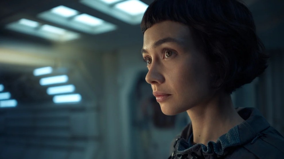 A woman with short dark hair and blunt bangs gazes ahead thoughtfully under soft, futuristic lighting.