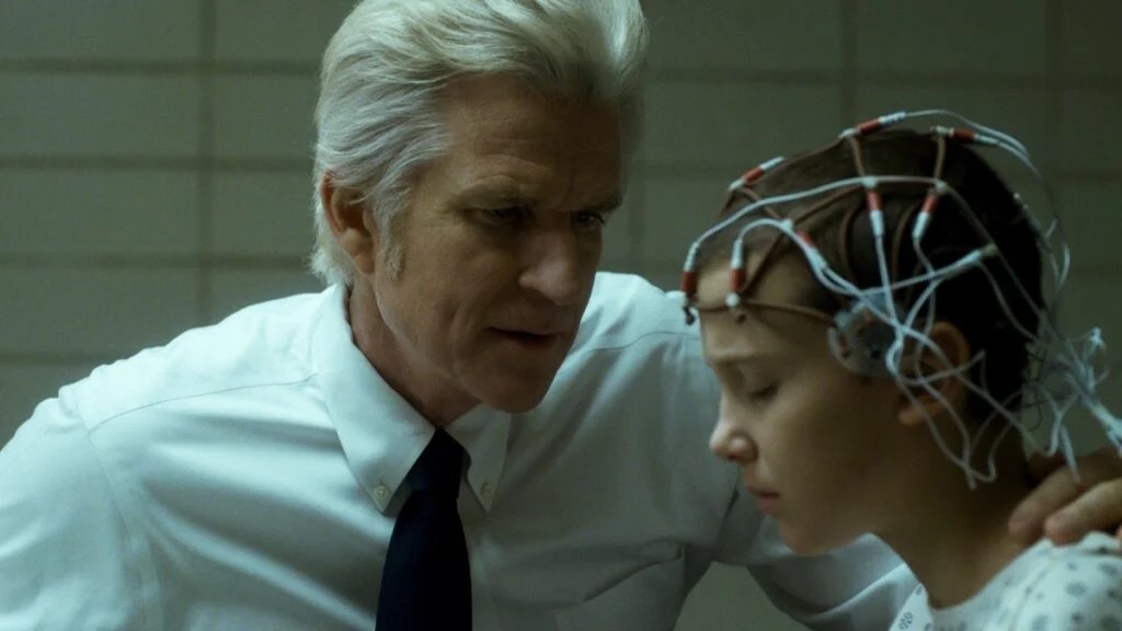A stern-looking scientist leans close to a young girl seated in a clinical lab setting. The girl wears a medical gown and an array of wires and electrodes on her shaved head. She looks tense and exhausted as he whispers something to her.
